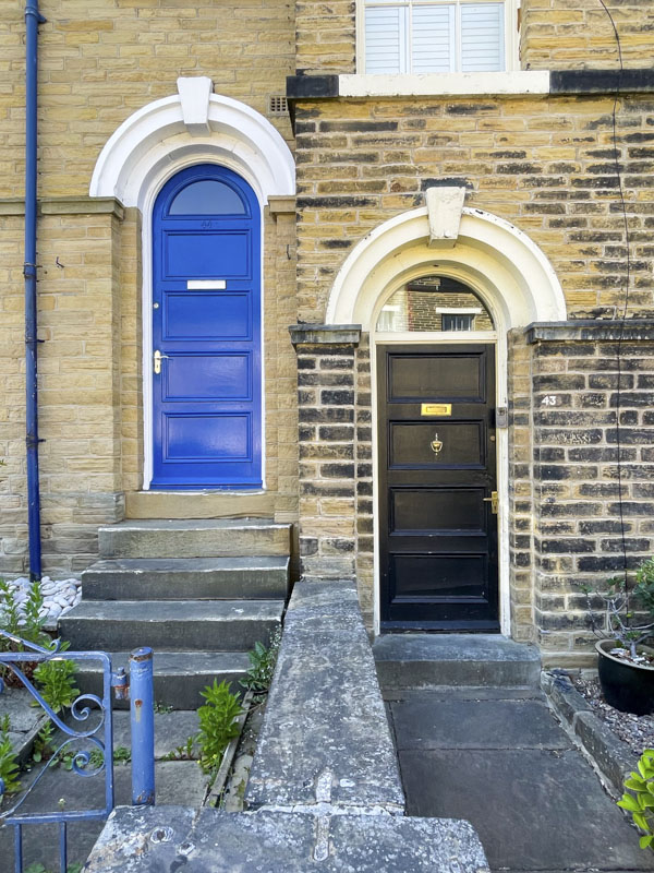 Raised blue door (two storey terrace) and slightly more fancy black door and surround (three storey end of terrace), George Street, Saltaire, West Yorkshire, England, May 2025